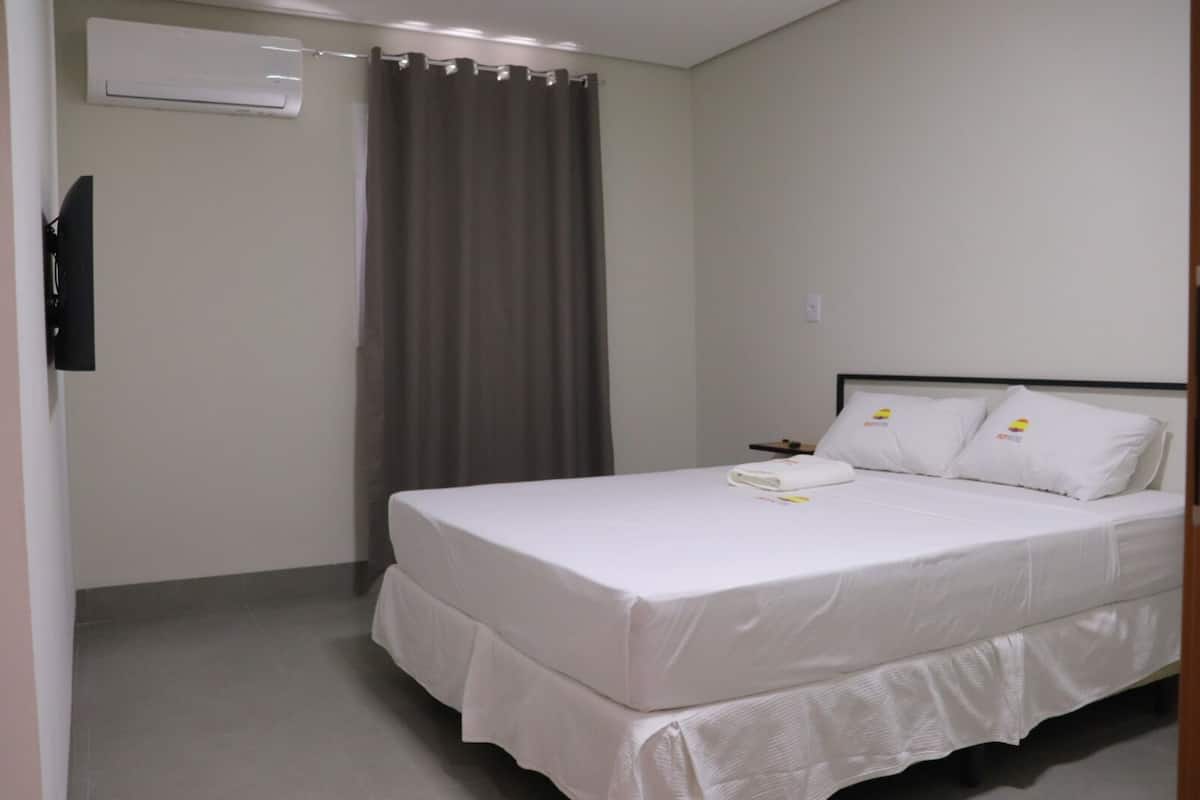 A neatly made bed with white linens and pillows, a flat-screen TV mounted on the wall, and a window with a curtain.