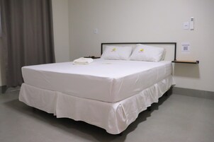 Una cama bien hecha con un colchón y almohadas blancos, una mesita de noche con una lámpara y un estante en la pared.