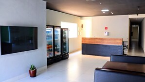 A modern lobby with a flat-screen TV, three beverage dispensers, a reception desk, and a potted plant. - Fly Hotel (Varzea Grande)