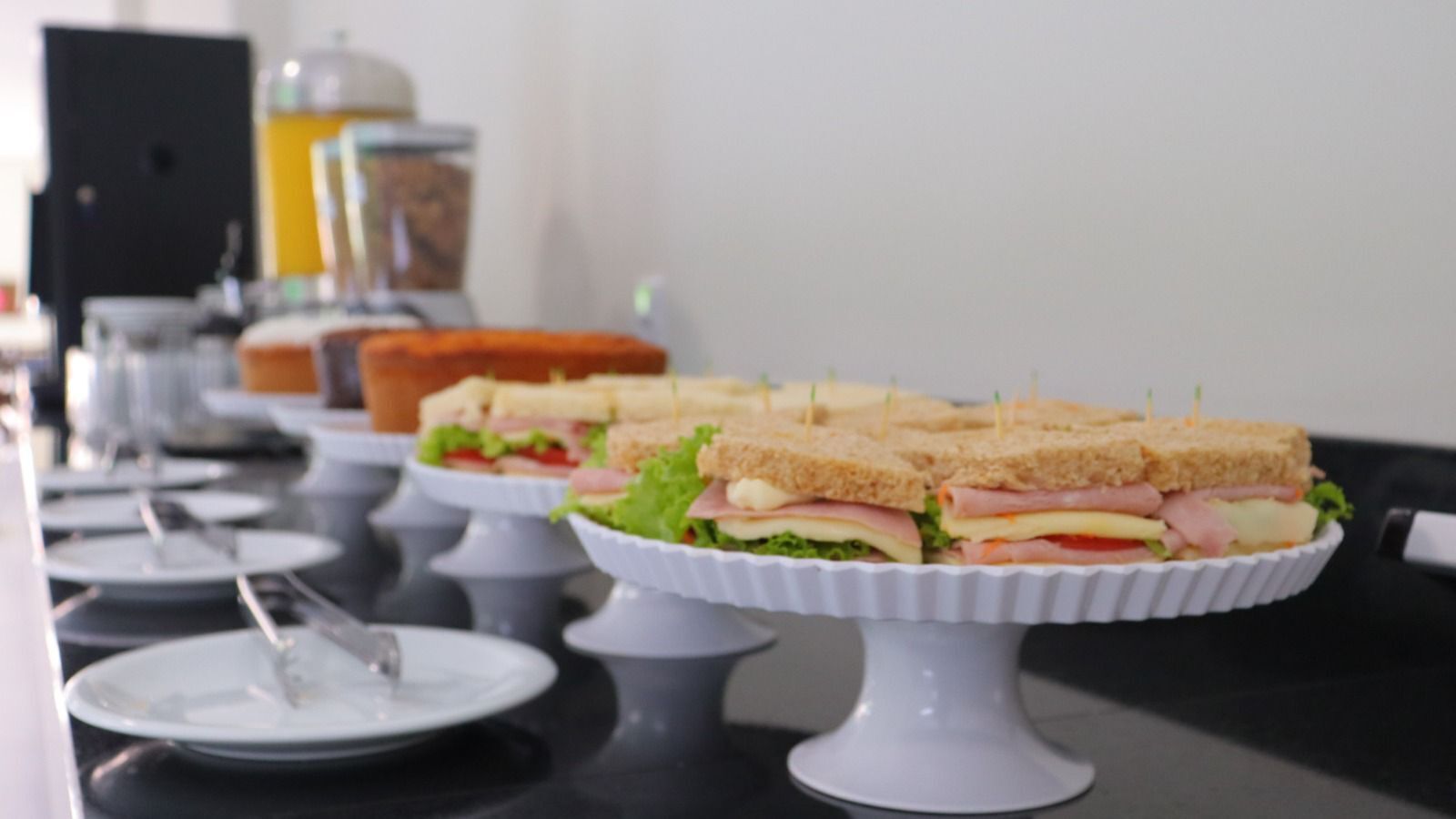 Disposição de buffet com sanduíches em suportes brancos, pratos e talheres sobre uma mesa.