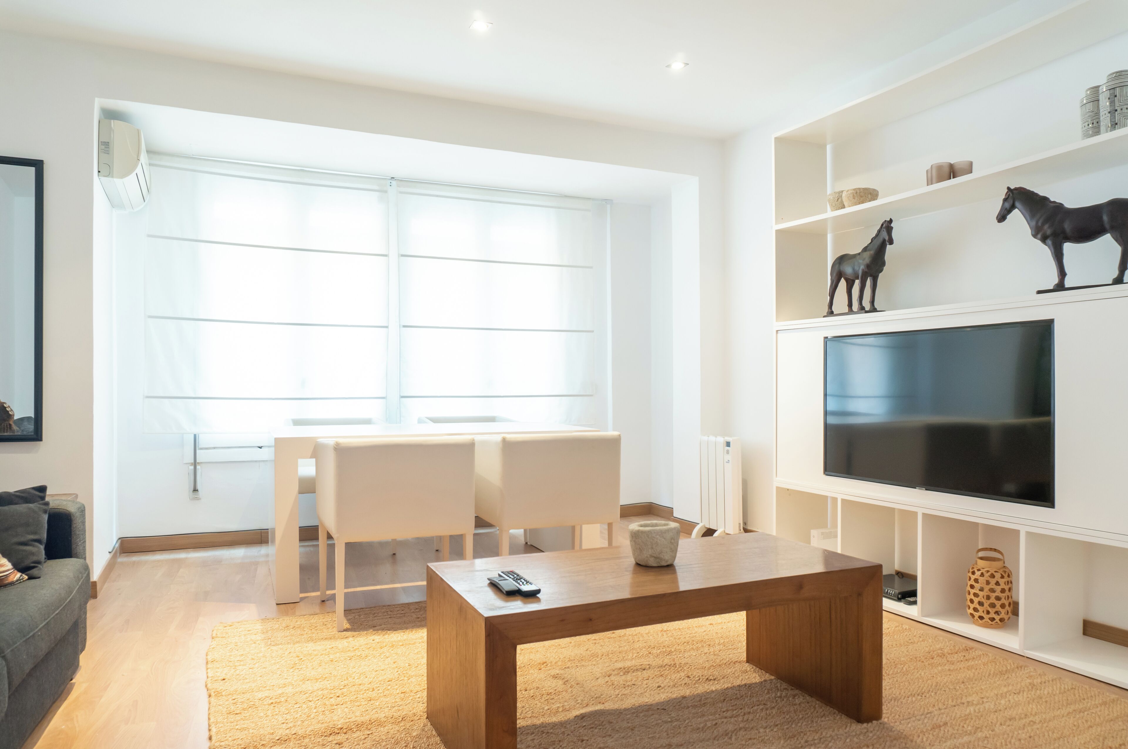 A modern living room with a large window, a dining area with two chairs, a wooden coffee table, and a mounted TV.