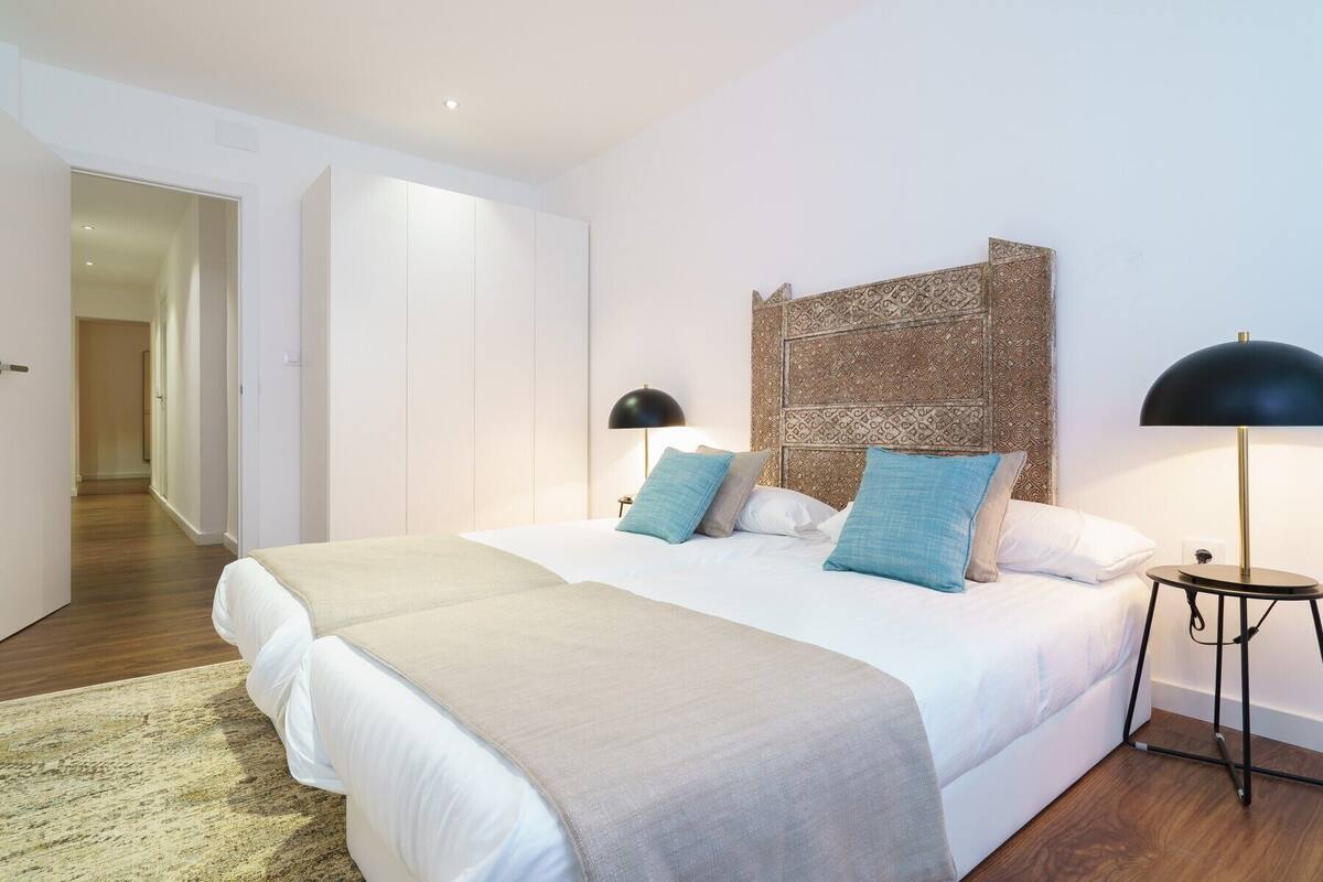 Een slaapkamer met een groot bed, twee nachtkastjes, een hoofdbord met een decoratief paneel, en een deur naar een andere kamer.