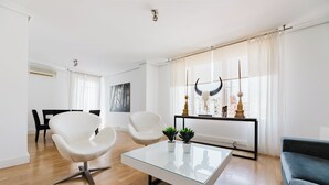 A modern living room with a glass coffee table, white armchairs, a sofa, and a dining area with black chairs. - Valencia Luxury - Boutique Center (Valencia)