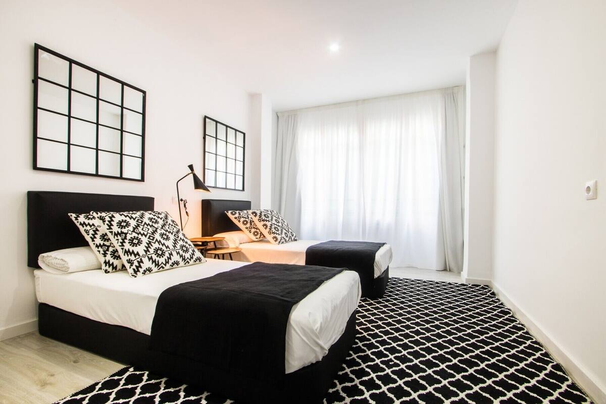 Een moderne hotelkamer met twee bedden, een groot raam met dunne gordijnen, een kamerbreed tapijt met dessin en een zwart-wit abstract schilderij aan de muur.