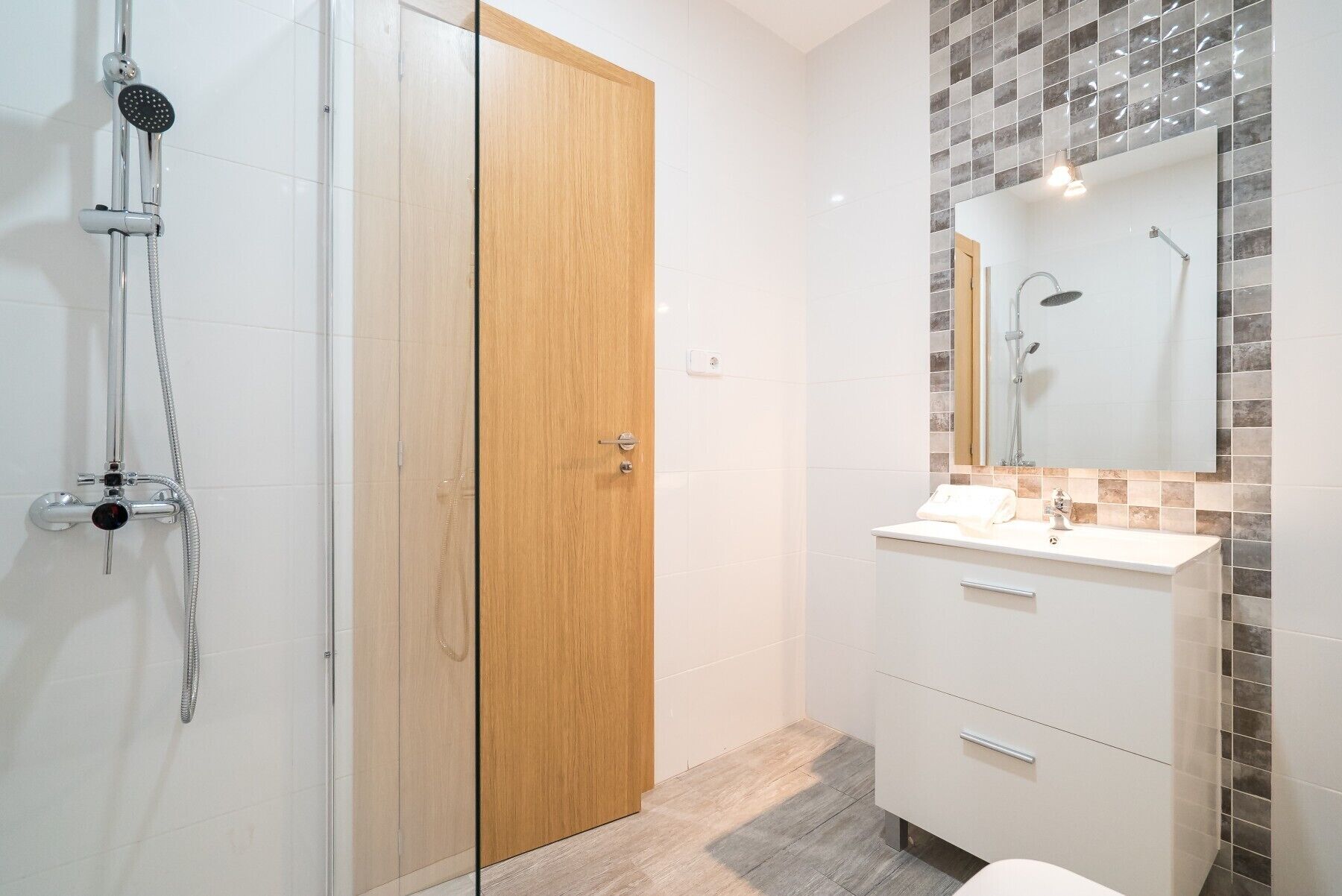 A modern bathroom with a glass shower enclosure, a wooden door, a vanity with a mirror, and a tiled backsplash.