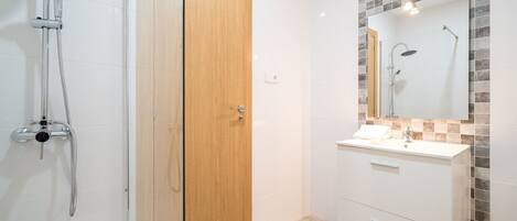 A modern bathroom with a glass shower enclosure, a wooden door, a vanity with a mirror, and a tiled backsplash.
