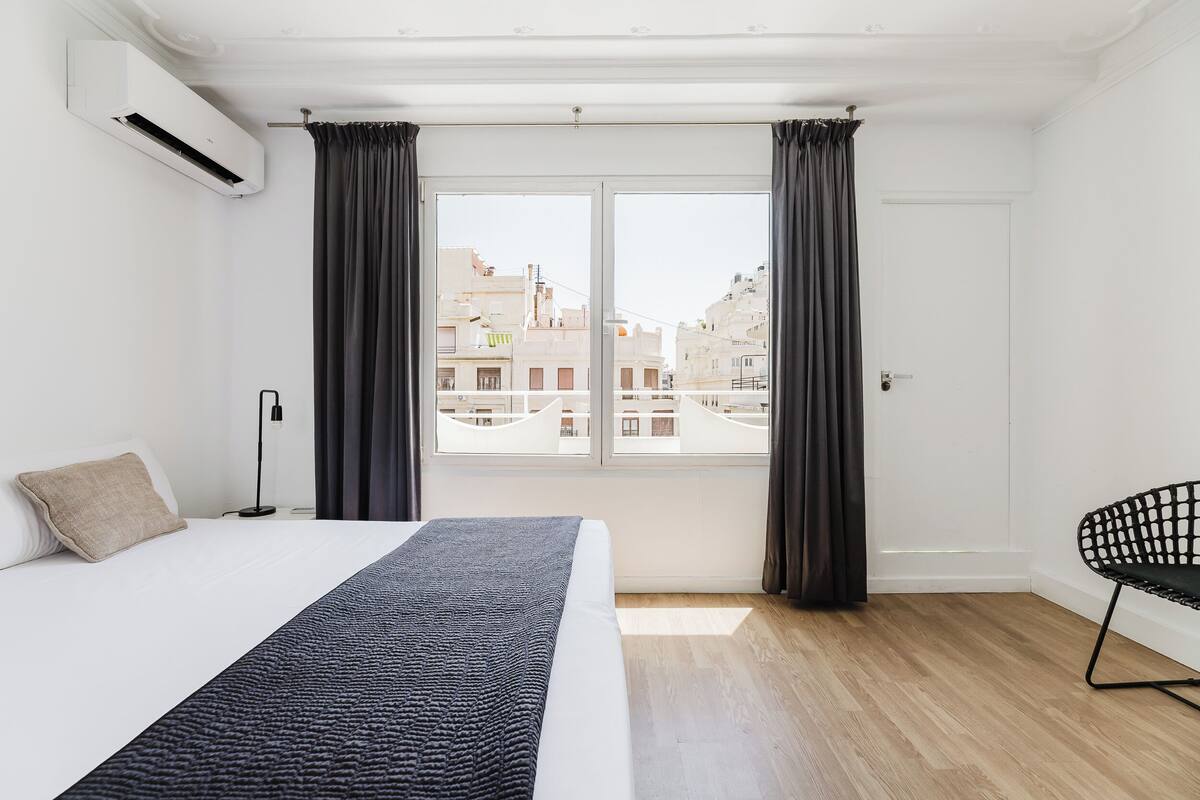 Een moderne slaapkamer met een groot bed, uitzicht op gebouwen door een raam en een zwarte stoel.