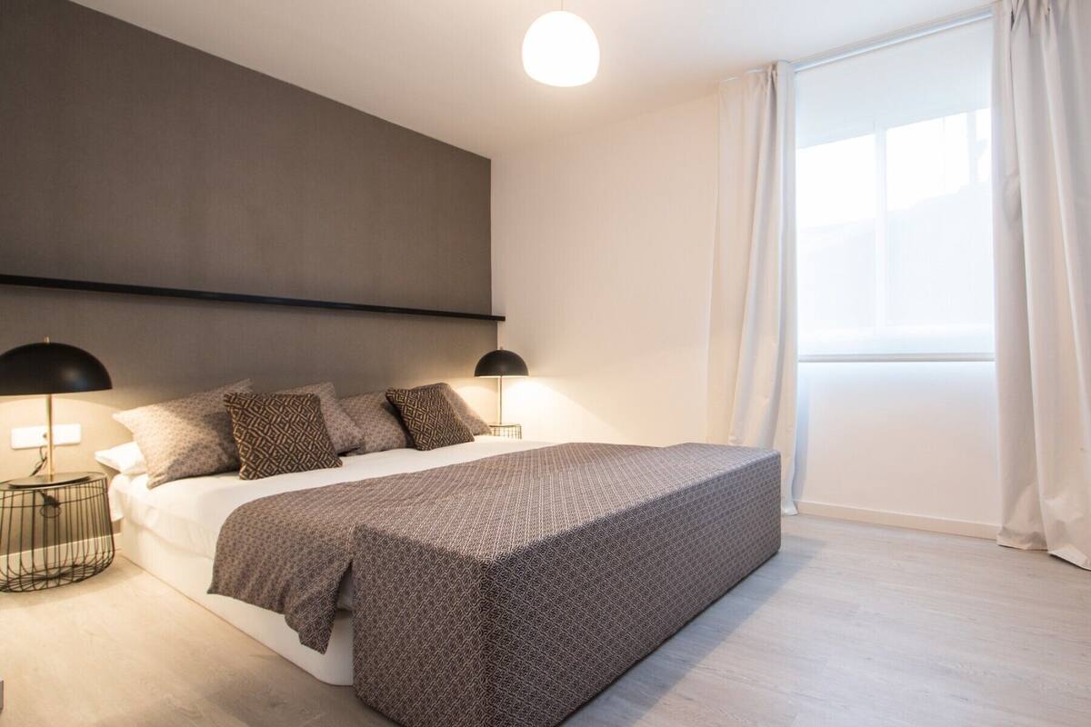 Een moderne slaapkamer met een groot bed, twee bedlampjes en een groot raam met gordijnen.