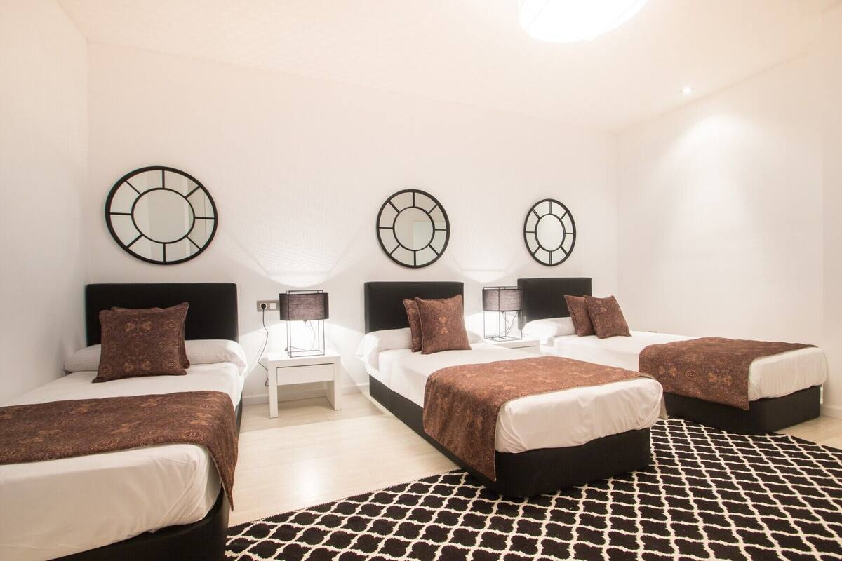 Een hotelkamer met drie bedden, een witte nachtkastje, een zwart-wit tapijt en drie spiegels aan de muur.