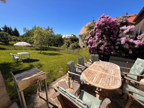 A backyard with a wooden table and chairs, a grill, and a pink flowering bush. - Cozy holiday apartment on a former farm in a quiet, green location (Hohnstein)