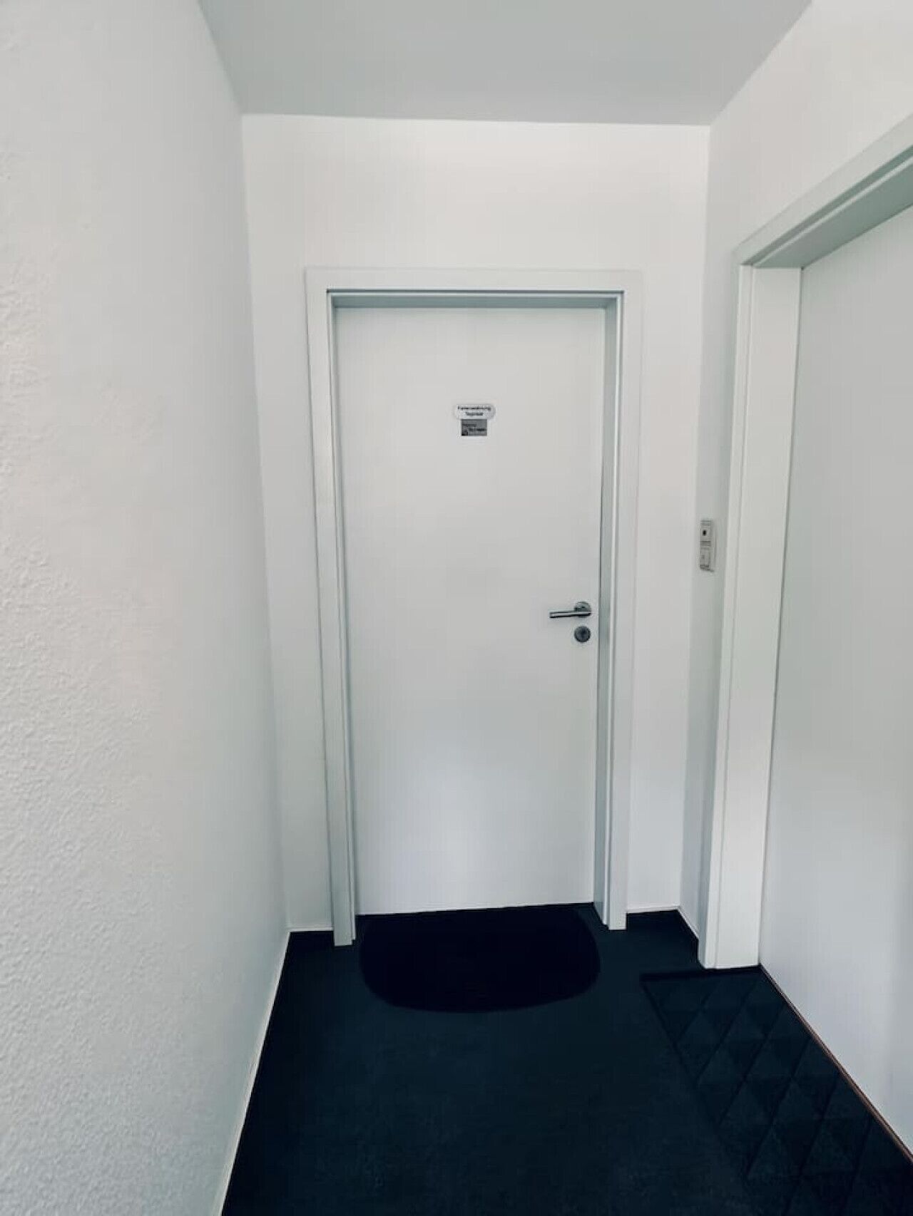 Ein schmaler Flur mit einer weißen Tür am Ende, einer schwarzen Matte am Eingang und einer weißen Wand an der Seite.