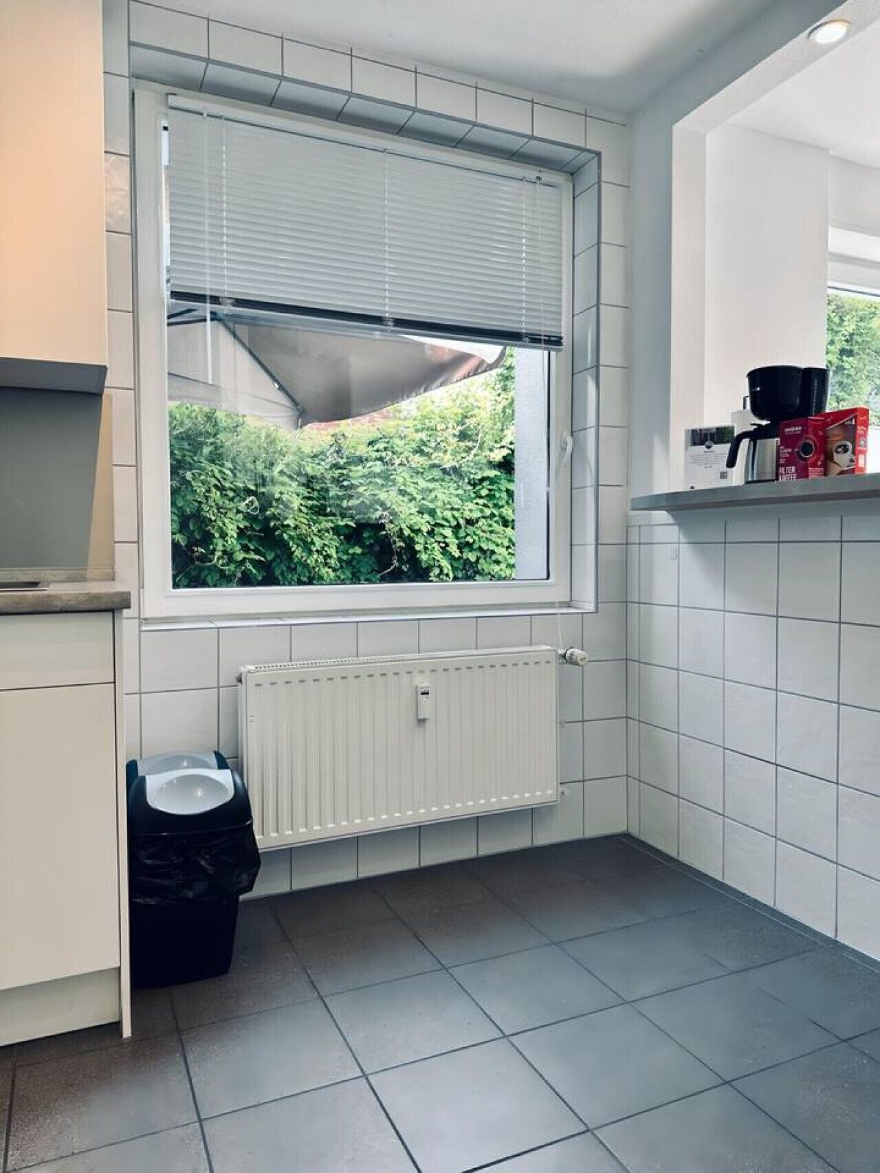 Une cuisine avec une fenêtre, des carreaux blancs, un radiateur et une poubelle.