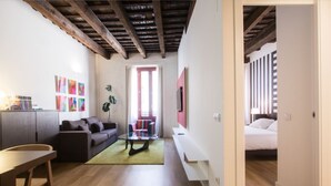Un salon moderne comprenant un canapé, une table basse et un coin repas. Une porte donne accès à une chambre à coucher où se trouve un lit.