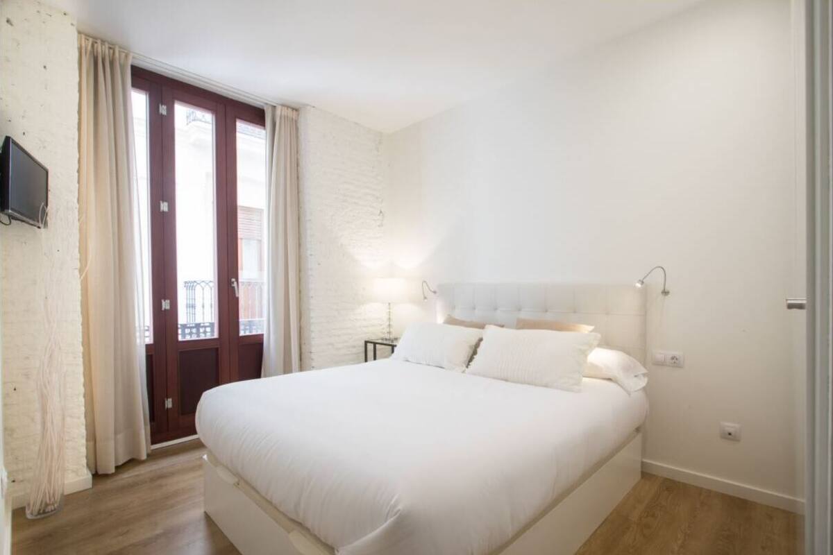 A bedroom with a large bed, a wooden door, a TV mounted on the wall, and a small bedside table with a lamp.