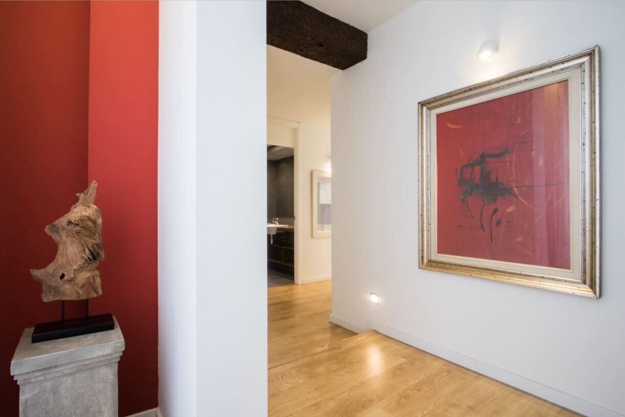 A hallway with a wooden sculpture on a pedestal, a framed abstract painting on the wall, and a glimpse of a kitchen area in the background.