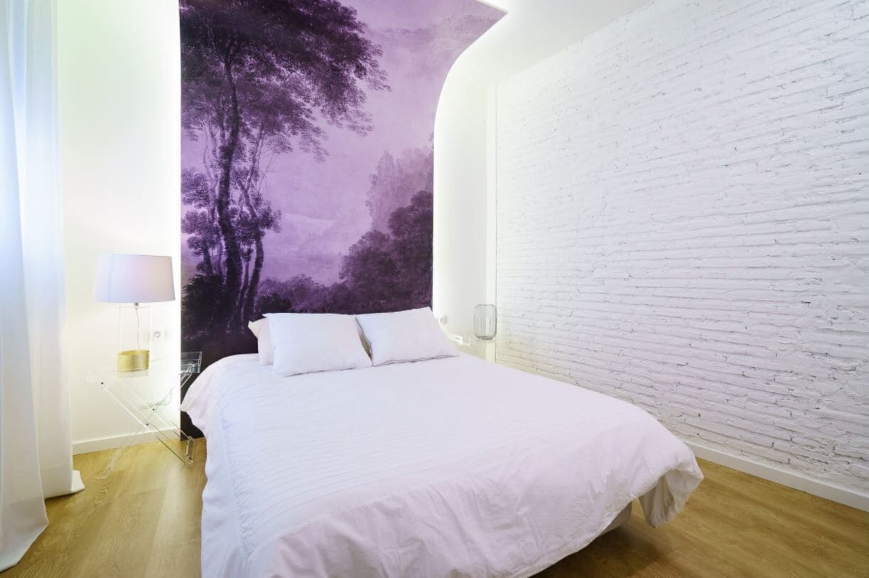 Un dormitorio con pared de ladrillo blanco, cama con ropa blanca y un mural de un árbol morado en la pared.