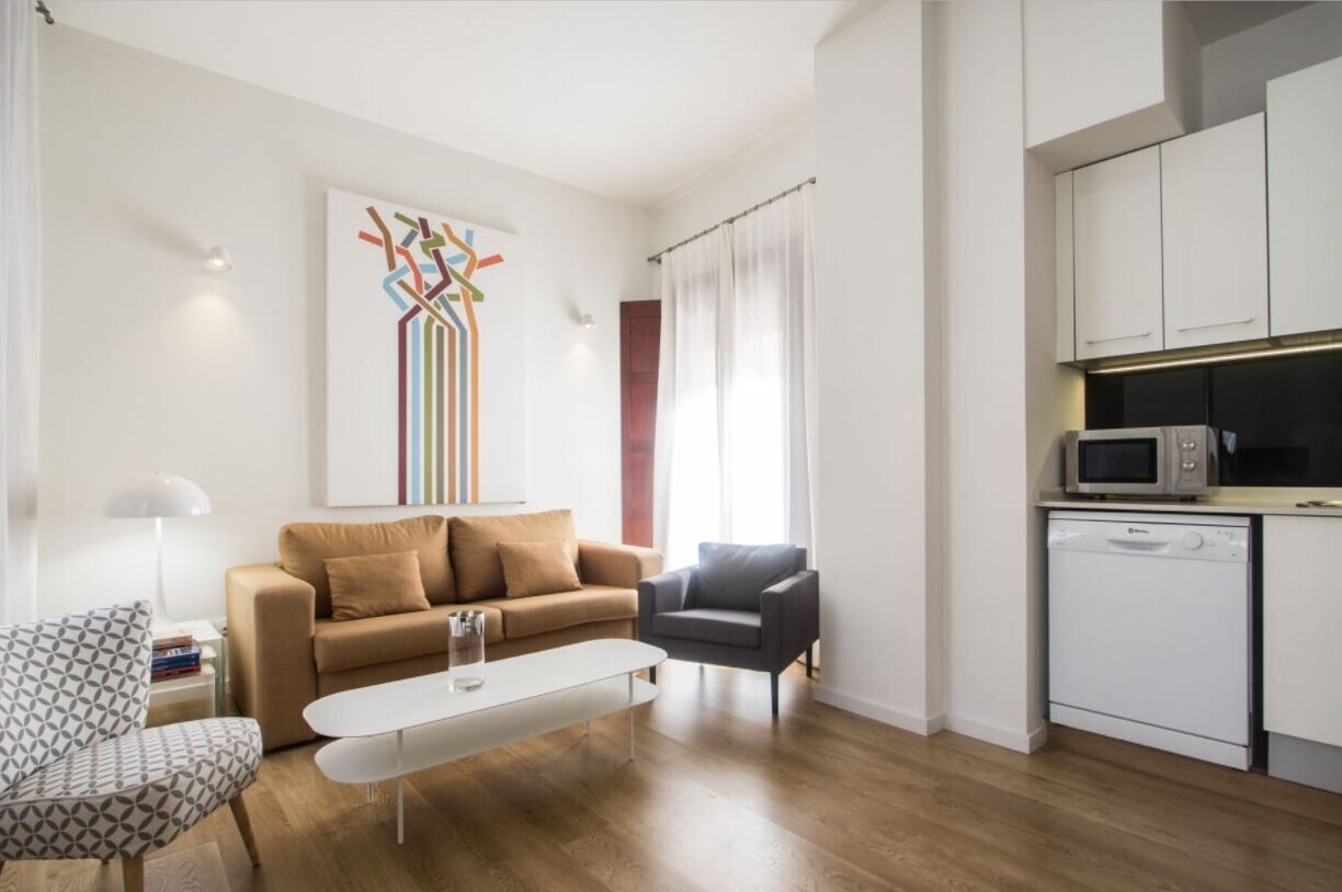 A modern living room with a sofa, a coffee table, a patterned armchair, and a kitchen area with a microwave and dishwasher.