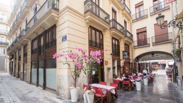 Un café en plein air avec des tables et des chaises, une composition florale et un bâtiment doté de balcons et d’une enseigne.