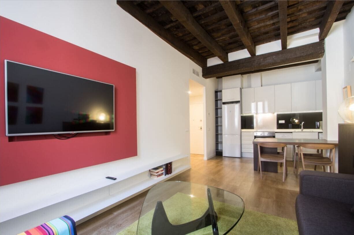 A modern living room with a flat-screen TV mounted on a red wall, a glass coffee table, and a sofa.