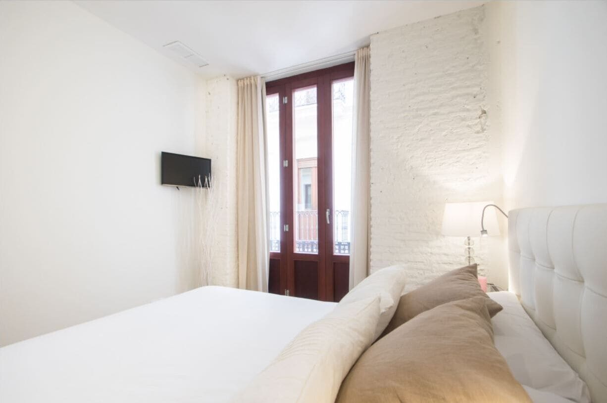 A hotel room with a bed, a flat-screen TV mounted on the wall, a window with curtains, and a balcony visible through the window.