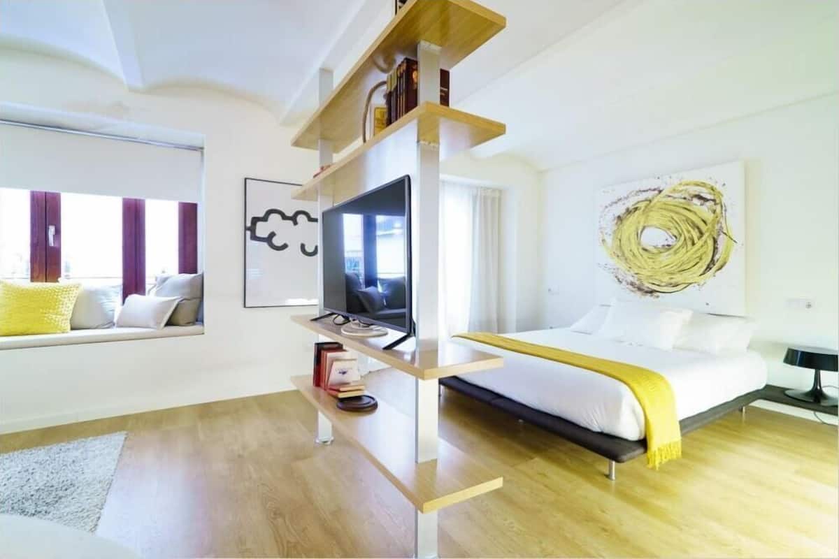 A modern bedroom with a bed, a TV on a stand, a window with curtains, and a wall art piece.