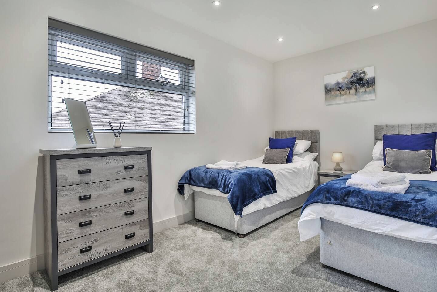 Une chambre avec deux lits, une commode, une fenêtre avec des stores et un tableau au mur.