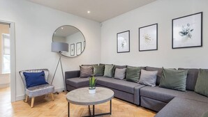 Een moderne woonkamer met een grijze bank, een ronde salontafel en een blauwe fauteuil. Er hangen kunstwerken in lijsten aan de muur en er staat een vloerlamp.