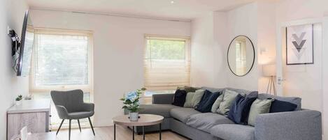 A modern living room with a grey sofa, a round coffee table, and a grey armchair. There is a small plant on the coffee table and a TV mounted on the wall.