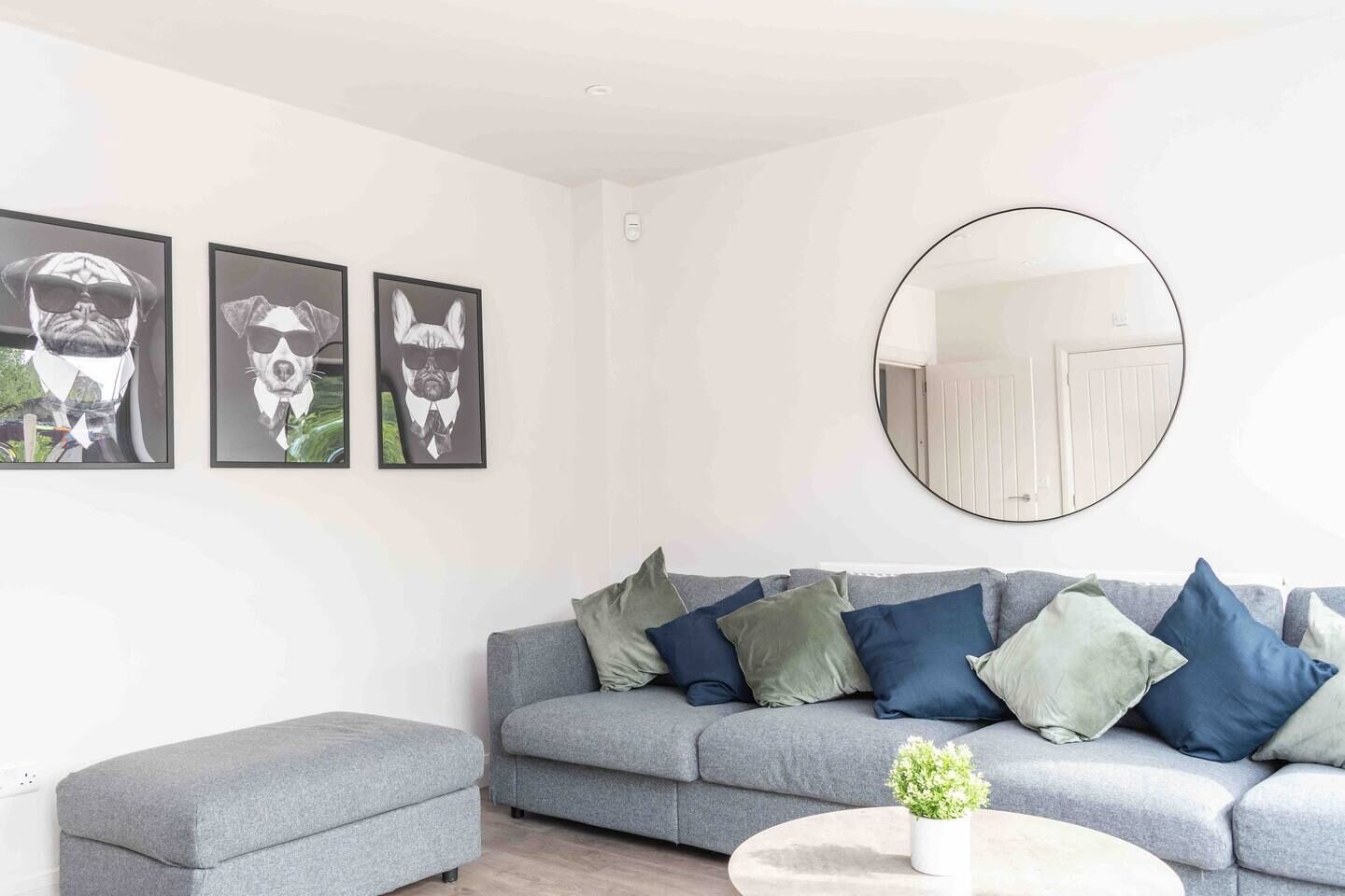 Un salon moderne avec un canapé gris, de nombreux coussins, un miroir rond et trois œuvres d’art encadrées accrochées au mur.