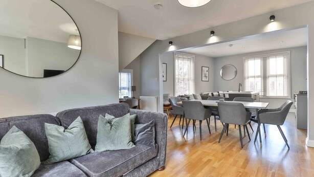 A modern living room with a grey sofa, dining table, and chairs. There is a large mirror on the wall and a staircase leading to another level. - Stylish & Spacious 3 Bed Apartment with Parking (Sheffield)