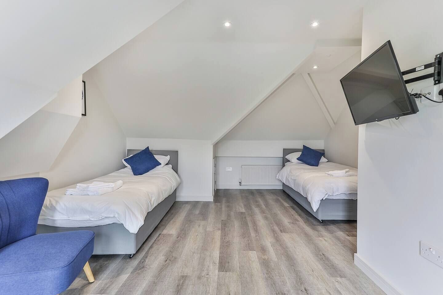 A modern, well-lit attic bedroom with two single beds, a blue chair, and a flat-screen TV mounted on the wall.