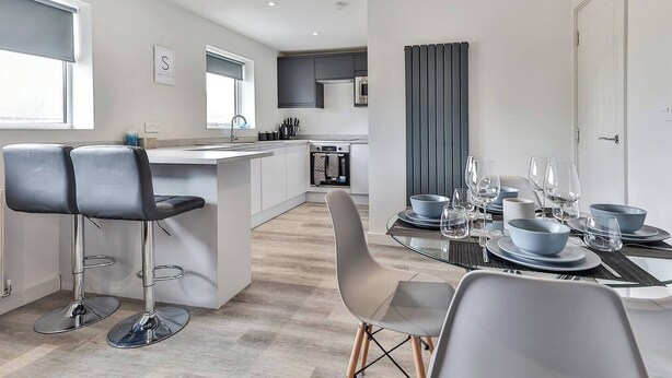 Una cocina moderna con una isla central, taburetes de bar y un comedor para cuatro con una mesa de vidrio y sillas.