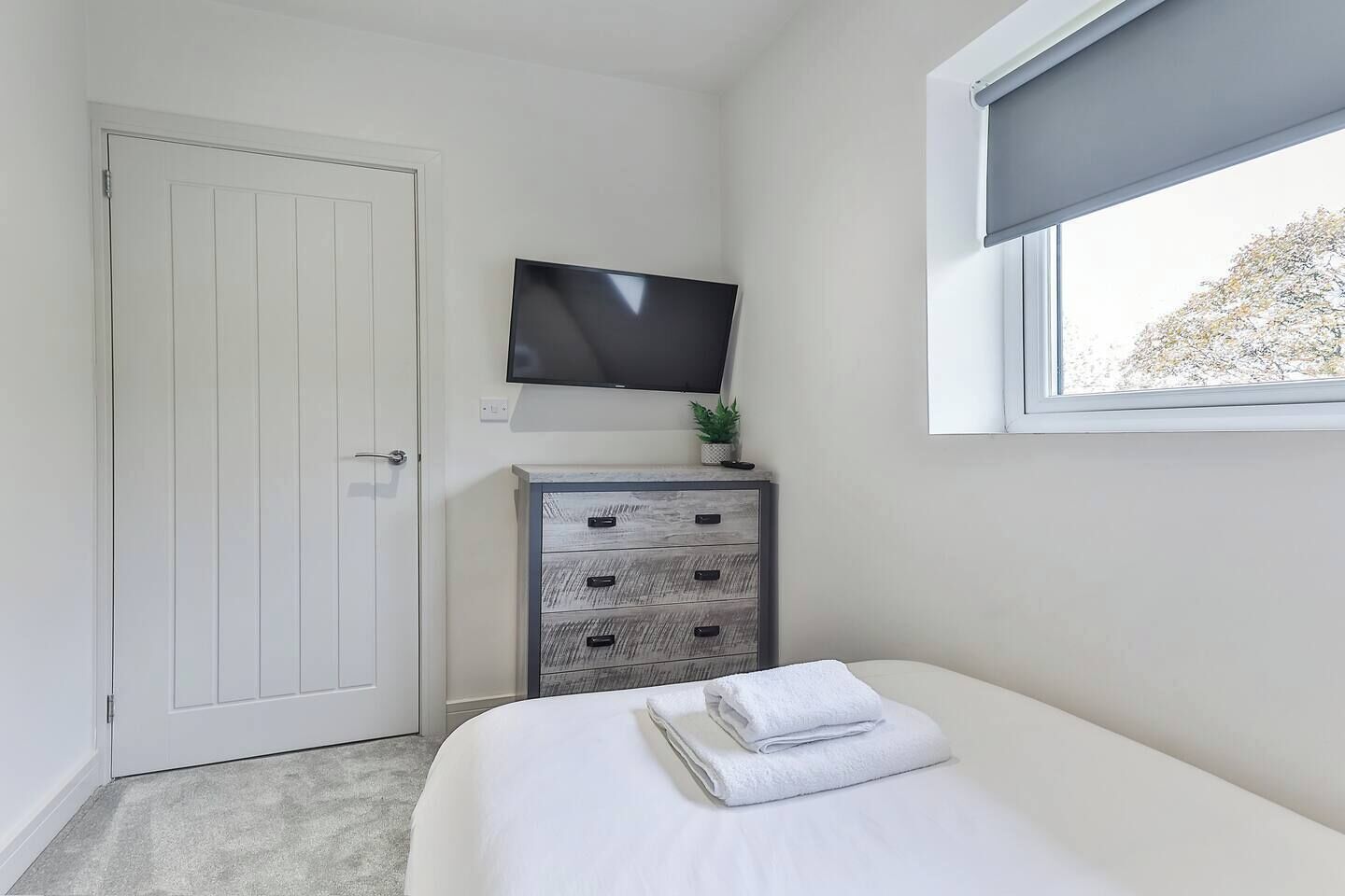 A bedroom with a bed, a dresser, a television mounted above the dresser, and a window with a view of trees.
