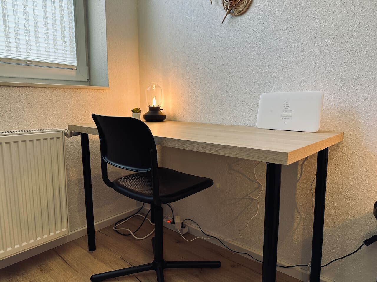 Ein kleiner Arbeitsbereich mit einem Holztisch, einem schwarzen Bürostuhl, einer weißen Lampe und einem weißen Gerät auf dem Tisch.