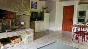 Un interior rústico con chimenea de piedra, un televisor sobre un mueble, una zona de comedor con sillas rojas y un piso de baldosas.