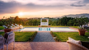 Uma área de piscina com espreguiçadeiras e um espaço de estar coberto por toldo, rodeada por vegetação e montanhas ao fundo.