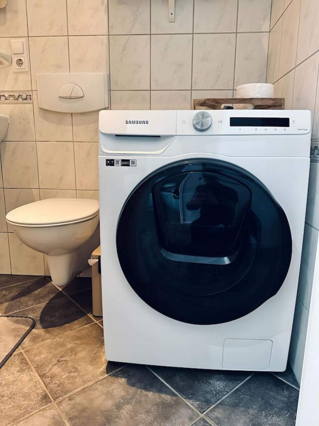 A Samsung front-loading washing machine in a bathroom with a toilet and tiled walls.
