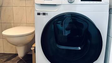 A Samsung front-loading washing machine in a bathroom with a toilet and tiled walls.