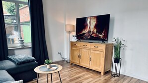 Un salon moderne avec un meuble en bois, une table basse ronde ornée d’une plante, un canapé et un téléviseur diffusant une image de cheminée.