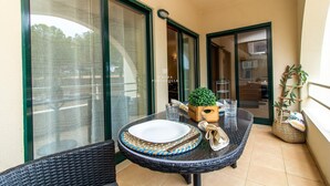 A balcony with a round table set for dining, a wicker chair, a potted plant, and a window with a view of the street. - Apartamento do Pinhal by D'Alma Portuguesa (Albufeira)
