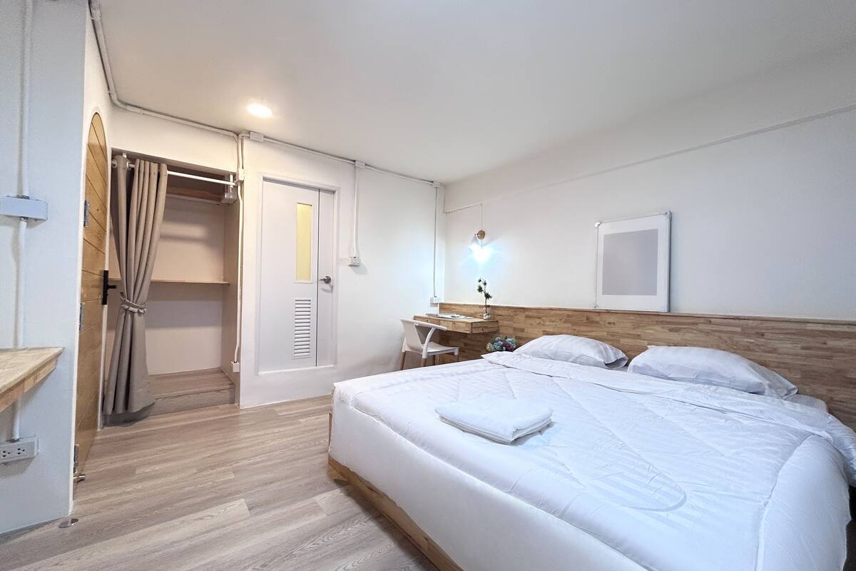 A hotel room with a wooden floor, a bed with white bedding, a desk with a chair, a nightstand, and a wall-mounted light.