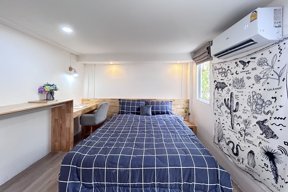 A bedroom with a large bed, a desk, and a chair. There is a window with a view of greenery.