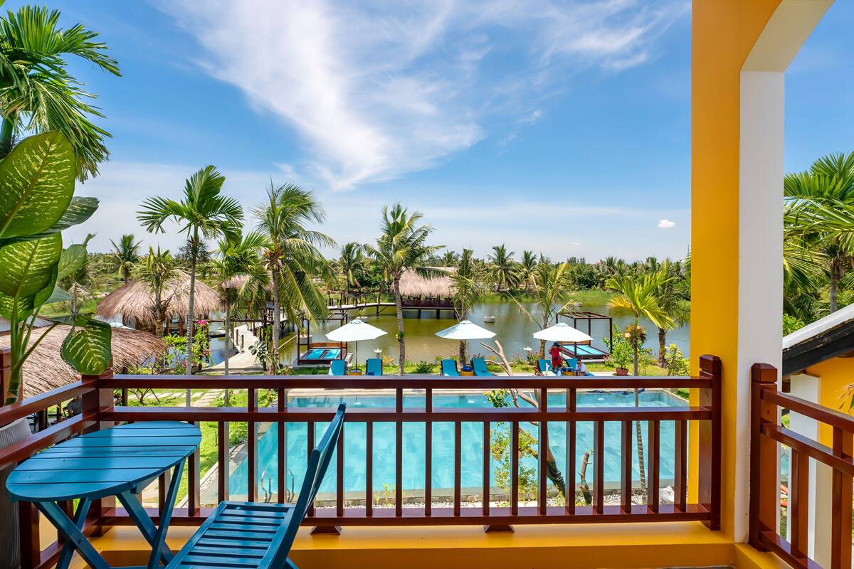 A balcony with a pool, lounge chairs, and umbrellas, overlooking a tropical resort area with palm trees and a thatched-roof hut.