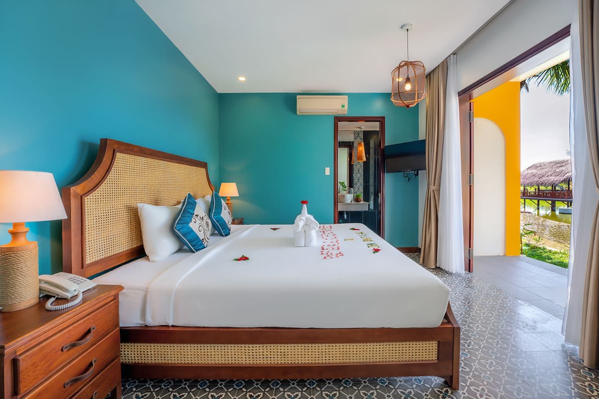 A hotel room with a large bed, a wooden headboard, a bedside table with a lamp, and a view of the outdoor area.