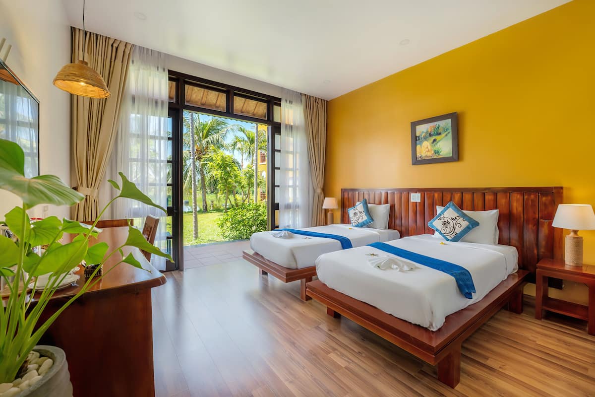 A hotel room with two beds, wooden flooring, a large window with curtains, a plant, and a painting on the wall.