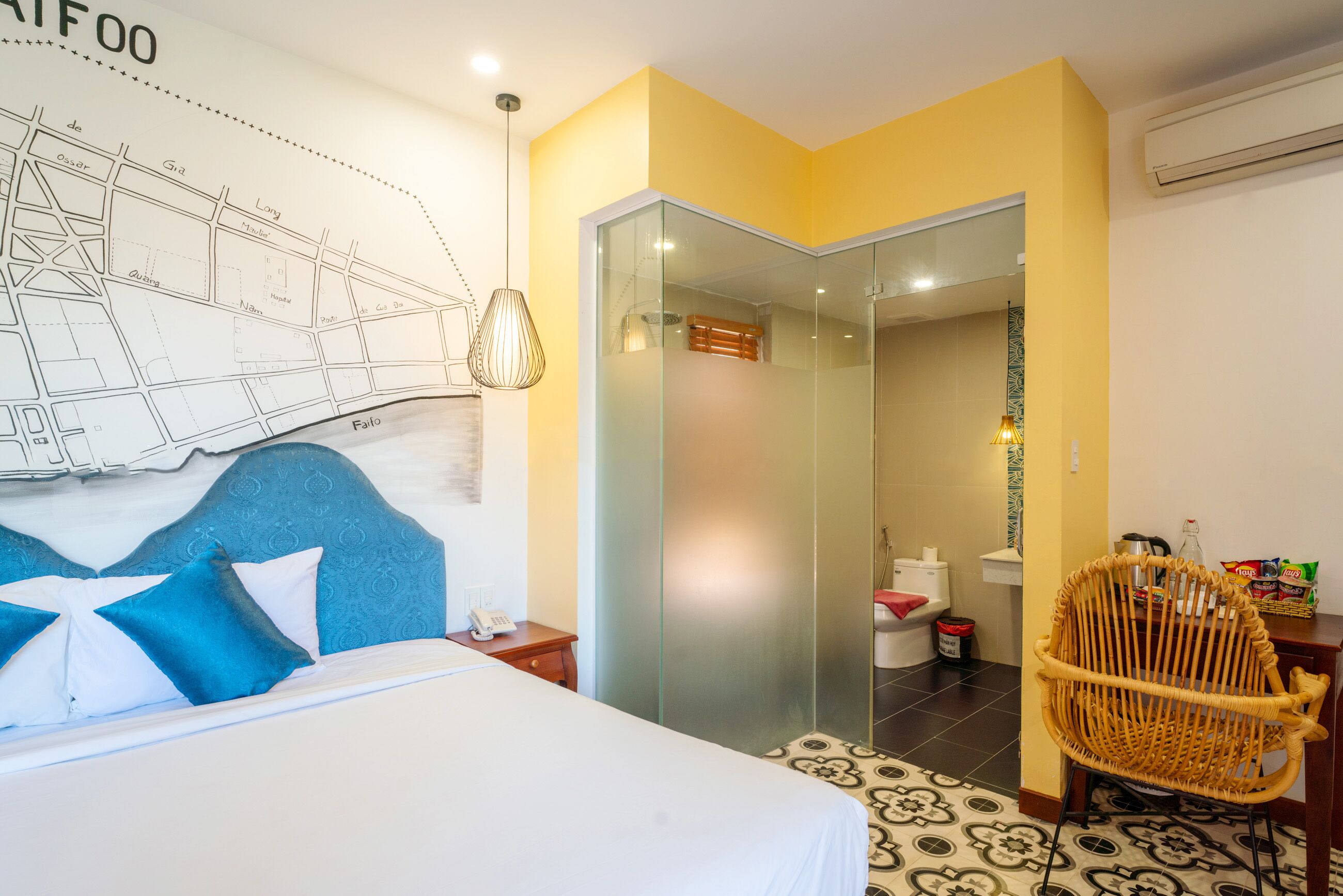 A hotel room with a bed, a map on the wall, a glass partition, a wicker chair, and a small table with snacks.