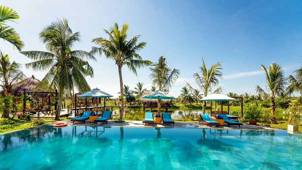 A clear blue pool with lounge chairs and umbrellas, surrounded by palm trees and a thatched-roof structure. - ZEN RETREAT HOI AN (Da Nang)