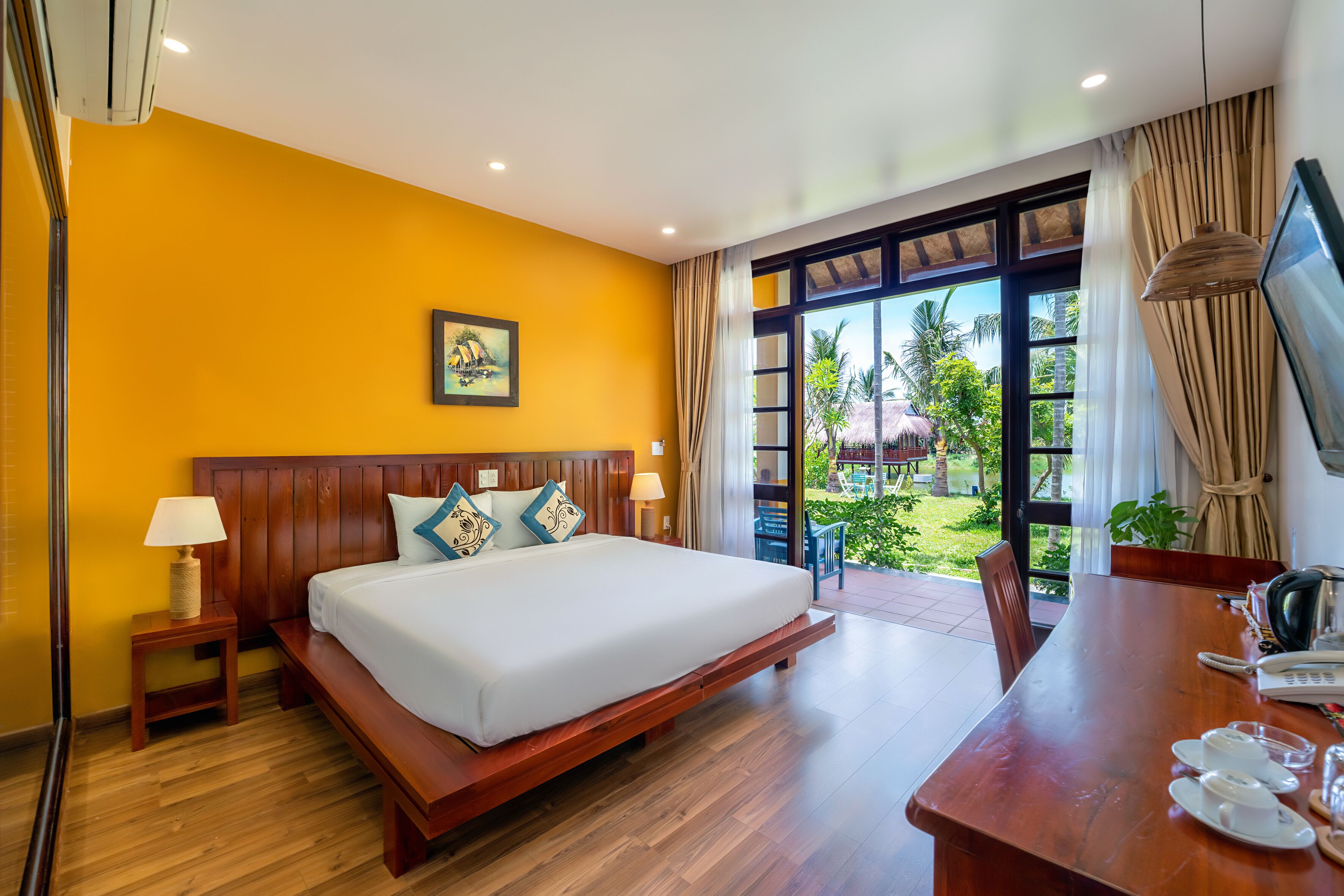 A hotel room with a large bed, wooden headboard, a dining table with chairs, a TV, and a view of the garden through a glass door.