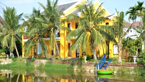 A yellow building with a reflection in the water, surrounded by palm trees. - ZEN RETREAT HOI AN (Da Nang)