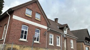 Uma casa de dois andares de tijolo com telhado inclinado, chaminé visível e janela com moldura branca.